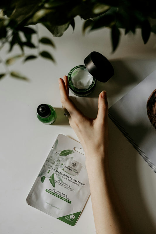 A hand reaching toward a jar of cream, a serum bottle, and a sheet mask